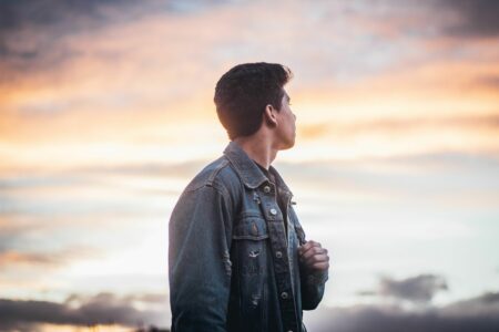 A young man in a denim jacket gazes at a vibrant sunset sky, capturing a contemplative moment.