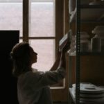 An artisan organizing ceramic plates on shelves against a distant window.