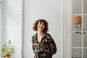 Happy woman in a floral top, standing indoors by a white wall and plant, smiling.